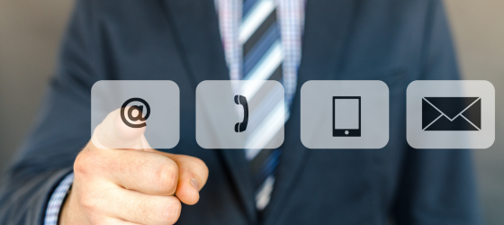 Uomo in abito professionale che punta il dito verso un'interfaccia digitale con icone di contatto. Le icone rappresentano un'email (@), un telefono fisso, uno smartphone e una busta da lettere